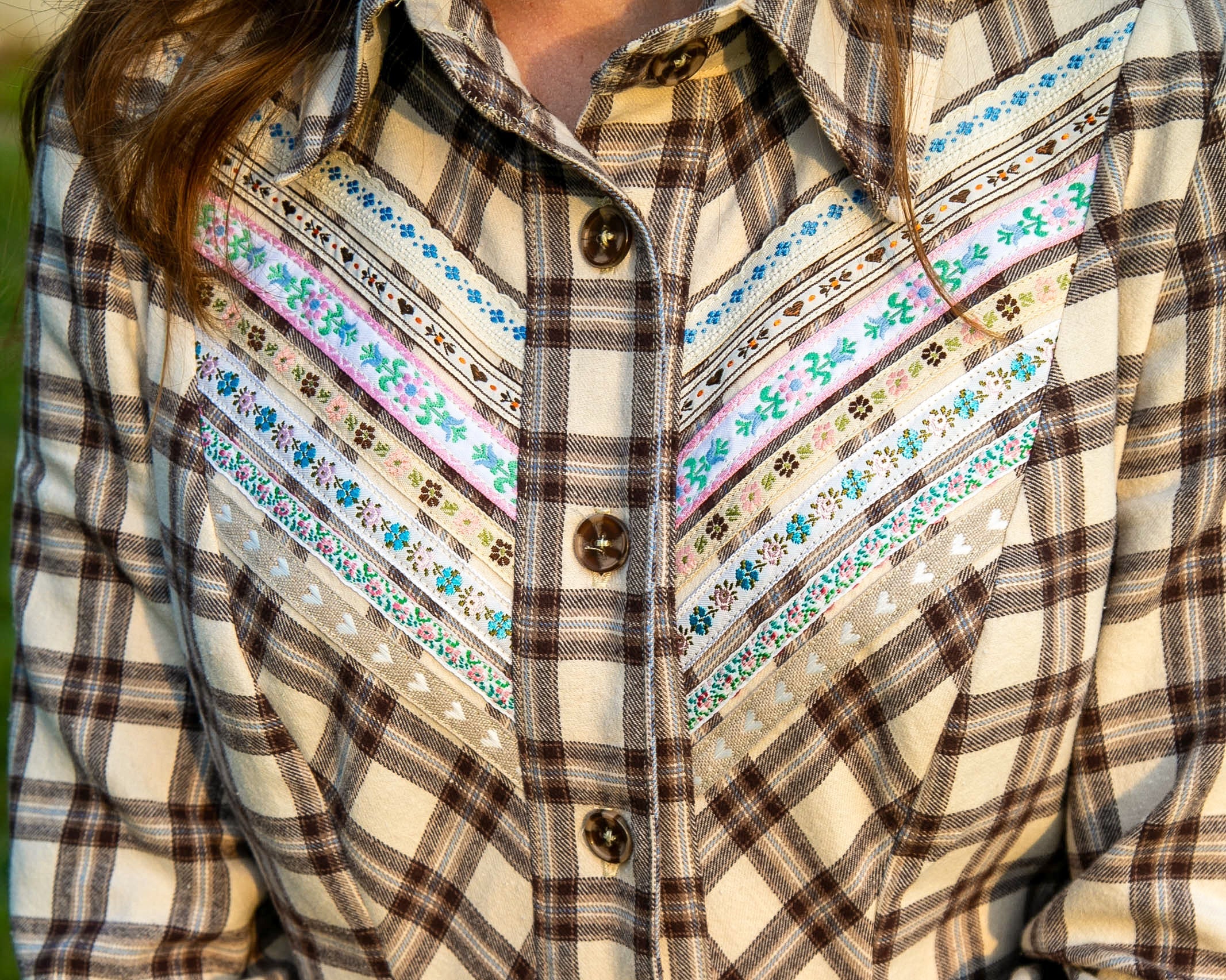 A close-up of a person wearing a long-sleeve plaid shirt dress in cream, brown, and gray, featuring embroidered yoke detailing with blue, pink, and green accents. The person is holding a brown leather-bound book.