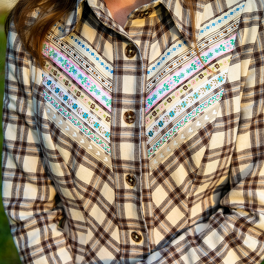 A close-up of a person wearing a long-sleeve plaid shirt dress in cream, brown, and gray, featuring embroidered yoke detailing with blue, pink, and green accents. The person is holding a brown leather-bound book.