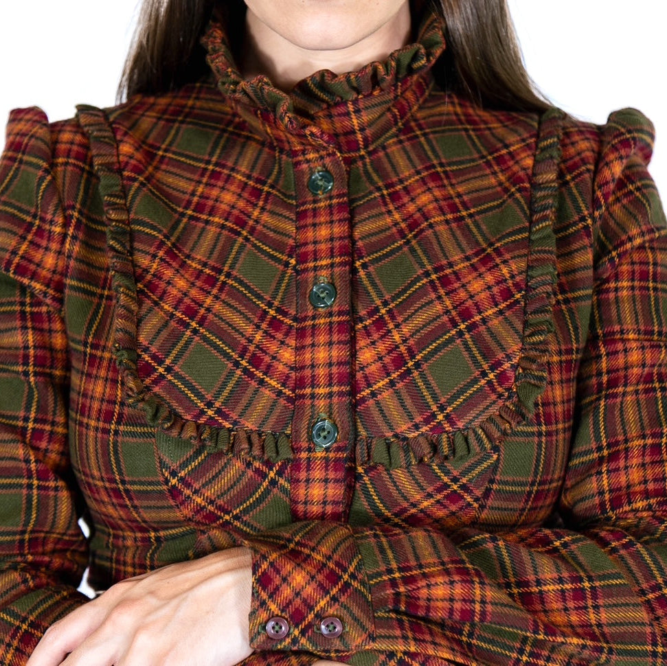 A close-up of a woman wearing a long-sleeve ruffled blouse with a green, red, and orange tartan plaid pattern, set against a plain white background.
