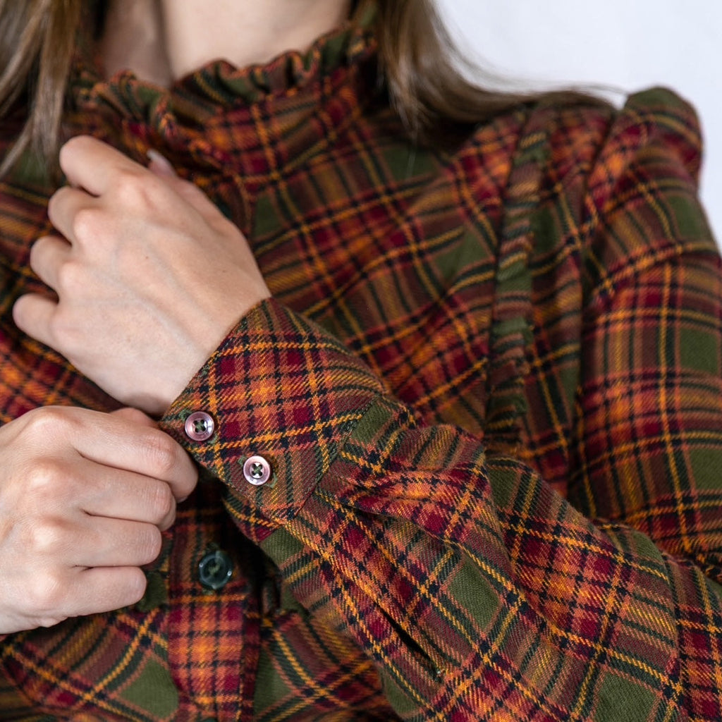 A close-up of a person wearing a long-sleeve plaid blouse with a ruffled high neck, adjusting the cuffs with buttons. The blouse features a green, rust red, and orange tartan pattern.