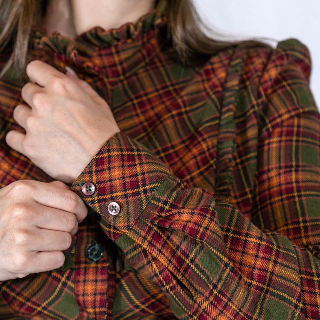 A close-up of a person wearing a long-sleeve plaid blouse with a ruffled high neck, adjusting the cuffs with buttons. The blouse features a green, rust red, and orange tartan pattern.