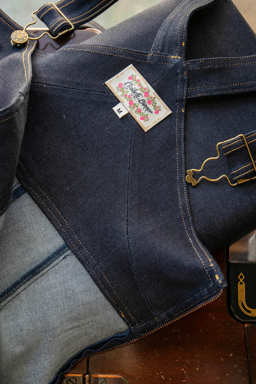 A dark indigo denim work apron with brass hardware and a striped inner lining, laid out on a wooden table next to a vintage sewing machine.
